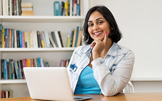 Latina woman facing camera in blue shirt and white jacket with laptop on table