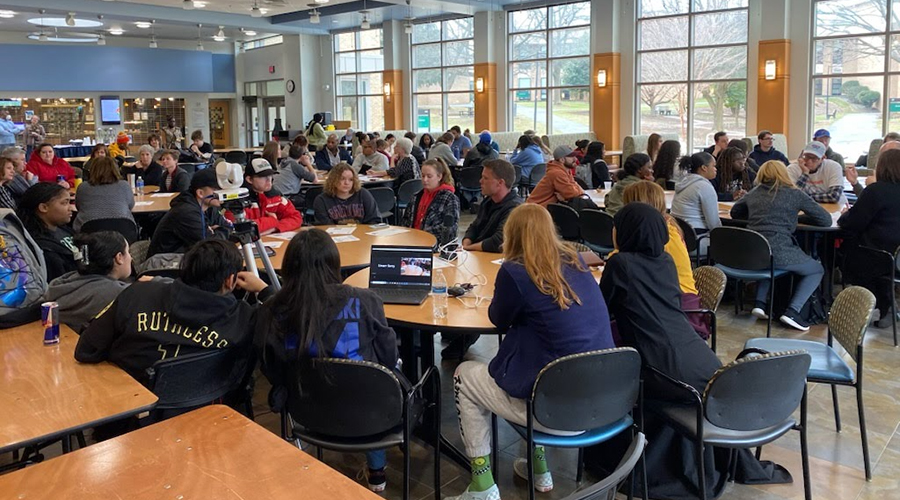 A large, window-filled room with groups of students and adults engaged in discussion at round tables. A camera on a tripod records a specific group gathered around an open laptop.