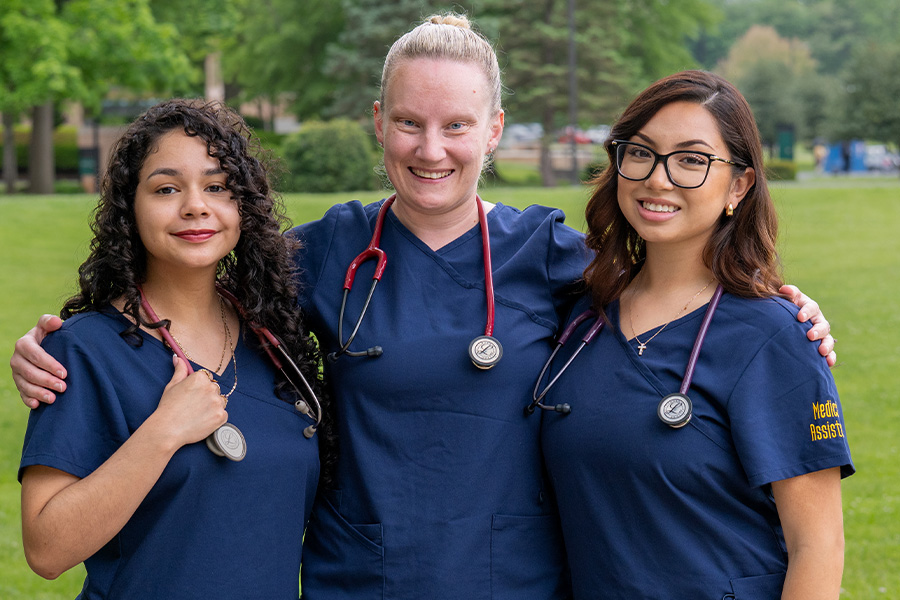 Three medical assistant students in navy scrubs with stethoscopes standing together outdoors on campus, smiling