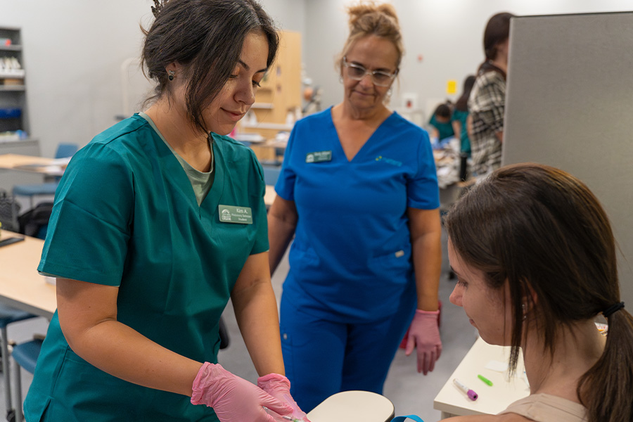 Phlebotomy student in teal scrubs preparing to draw blood from a classmate while an instructor observes