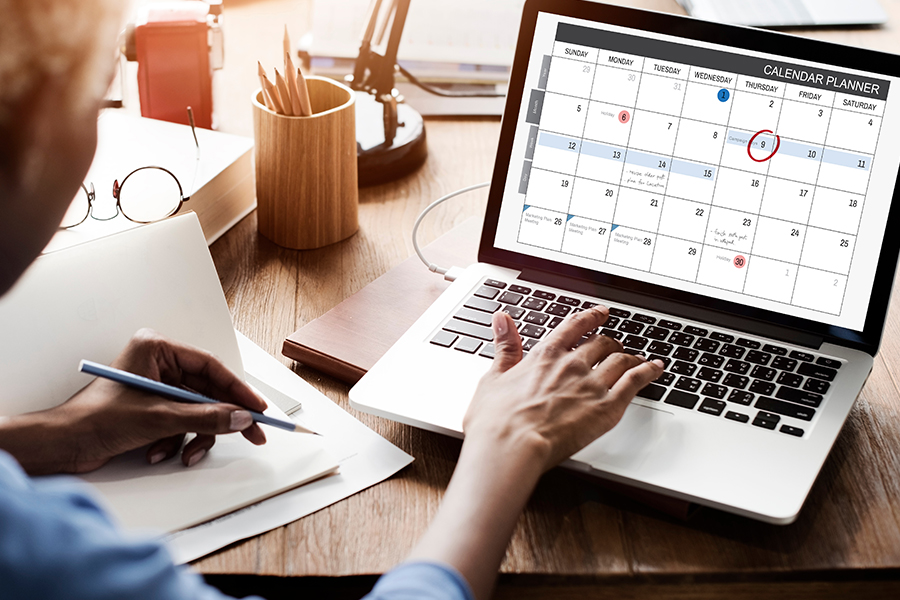 Person typing on a laptop displaying a calendar planner. A red circle highlights a date. Nearby, a notepad and glasses suggest planning or organizing tasks.