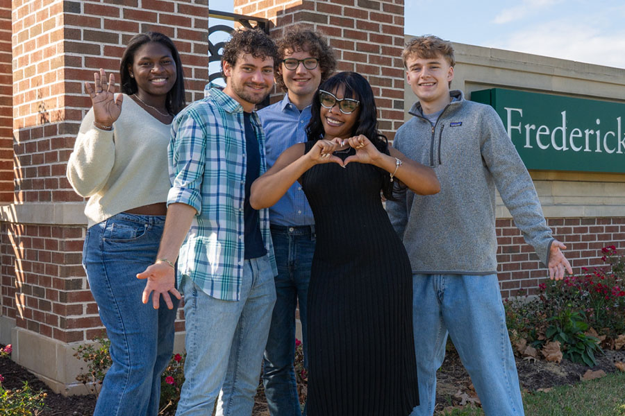 Five FCC students posing together outside near the Frederick Community College sign, smiling and waving.