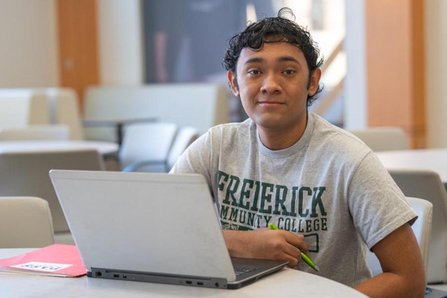 A student wearing a grey Frederick Community College t-shirt smiles while working on a silver laptop in a bright, open campus common area.
