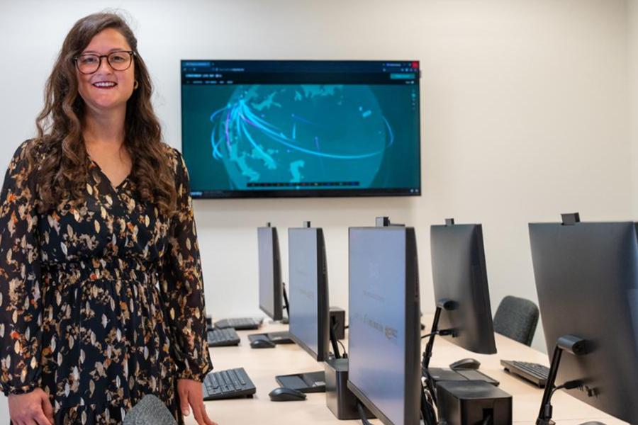 A smiling professional woman stands in a modern computer lab featuring rows of workstations and a large wall-mounted screen displaying a digital global network graphic.