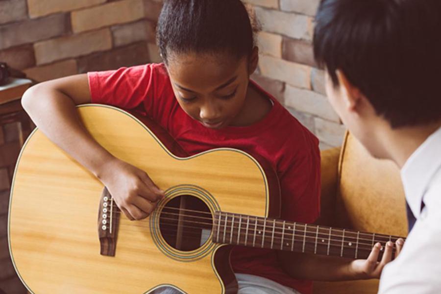 A young music student in a red shirt receives a private guitar lesson, focusing on finger placement on the fretboard.